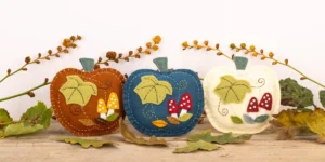 3 pumpkin felt ornaments in rust, teal and cream, on a shelf with autumn leaves