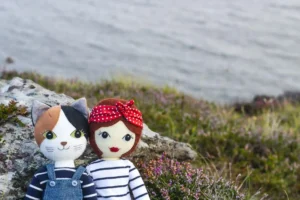 Tilly doll and Puffin the Cat doll sitting against a rock with flowering heather and the sea in the background.