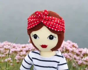 A felt doll wearing a navy and white striped top and a red and white spotted scarf around her hair, with the sea and sea pink flowers in the background.