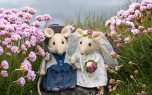 A felt mouse bride in a rose patterned dress and groom in top hat and tails, standing among sea pink flowers with the sea in the background.