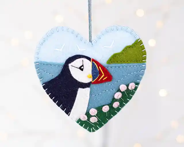 A felt heart ornament appliqued and embroidered with a puffin's head and shoulders, sea pink flowers and the sea and coastline in the background