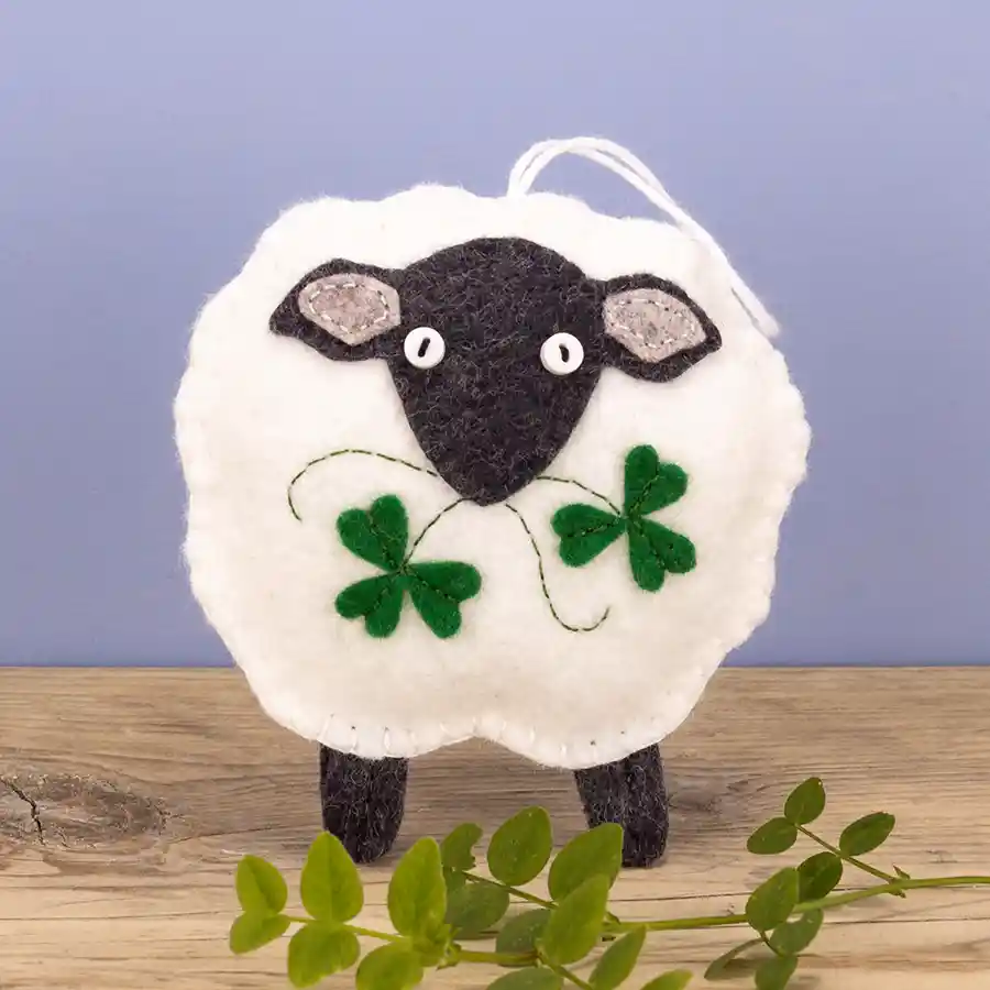 A white felt sheep ornament with dark grey head and legs, eating shamrock leaves and stems.