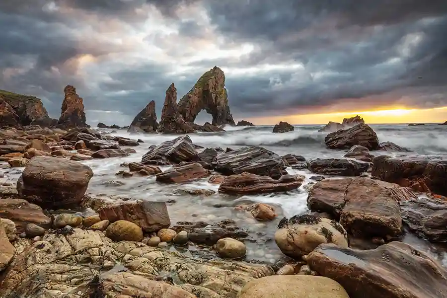 Photograph of a group of rocks including the Crohy Head sea arch, with a dramatic sky and waves.
