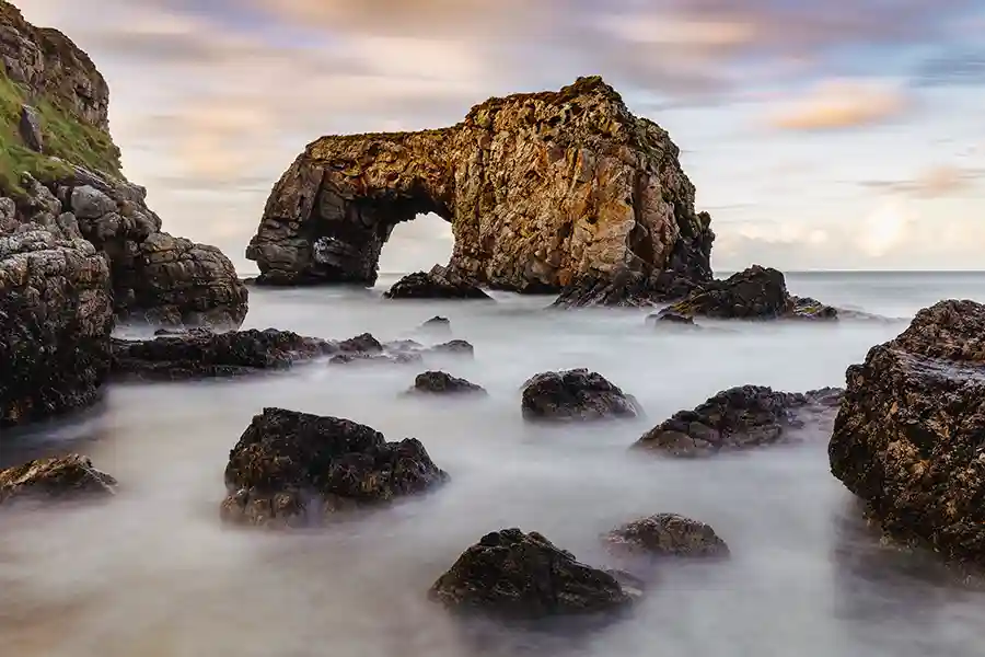 Photograph of a dramatic rock arch among a group of rocks, with a misty effect on the sea created by long exposure.