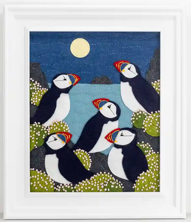 An embroidered felt applique showing five puffins looking up at the full moon, surrounded by clumps of sea thrift flowers. Framed in a wide white wooden frame.