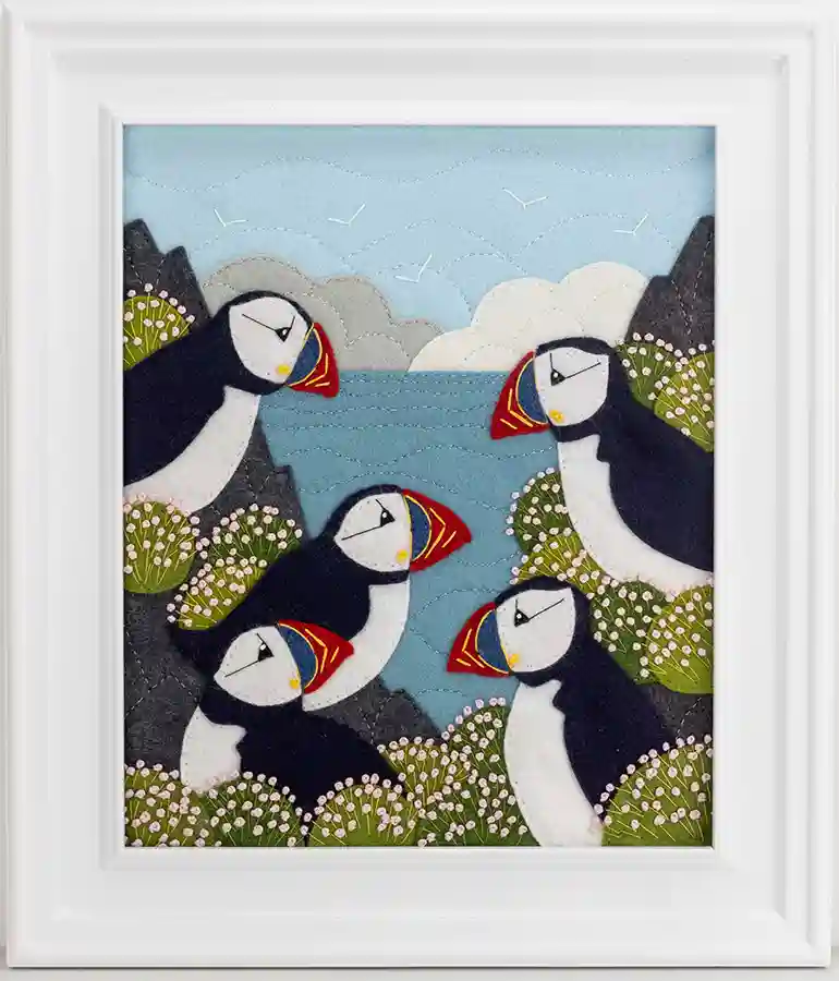 An embroidered felt applique showing five puffins among the rocks, surrounded by clumps of sea thrift flowers. The sea and cloudy sky are in the background. Framed in a wide, white wood frame.