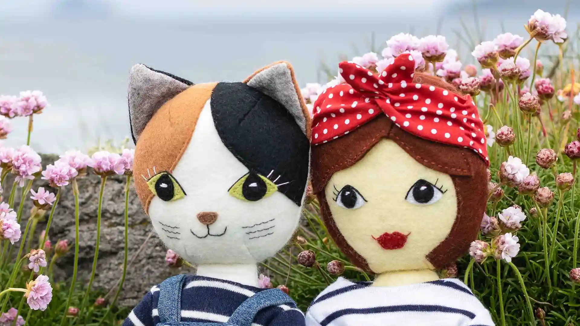 Tilly and Puffin, a felt cat and felt girl doll, sitting on a clifftop among the sea pink flowers.