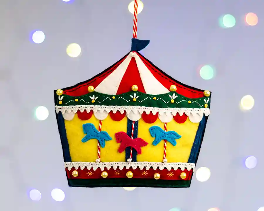 Carousel felt ornament with red and white striped canopy, red, green, yellow and blue decoration, three little felt horses with striped poles, and glass beads to represent strings of lights.