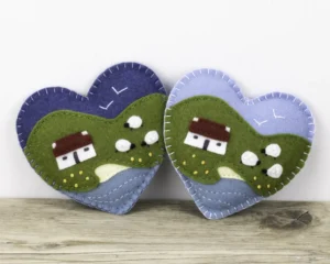 Two felt heart ornaments with appliqued and embroidered cottage and sheep on a background of hills with a beach and the sea in the foreground. Flying gulls are stitched on the sky. One has a light blue sky and one dark blue.