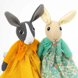 Two felt rabbit toys, one made in light brown felt wearing a green floral Liberty print dress and green felt jacket, and one in grey and white wearing an orange and white polka dot dress with an orange felt jacket.