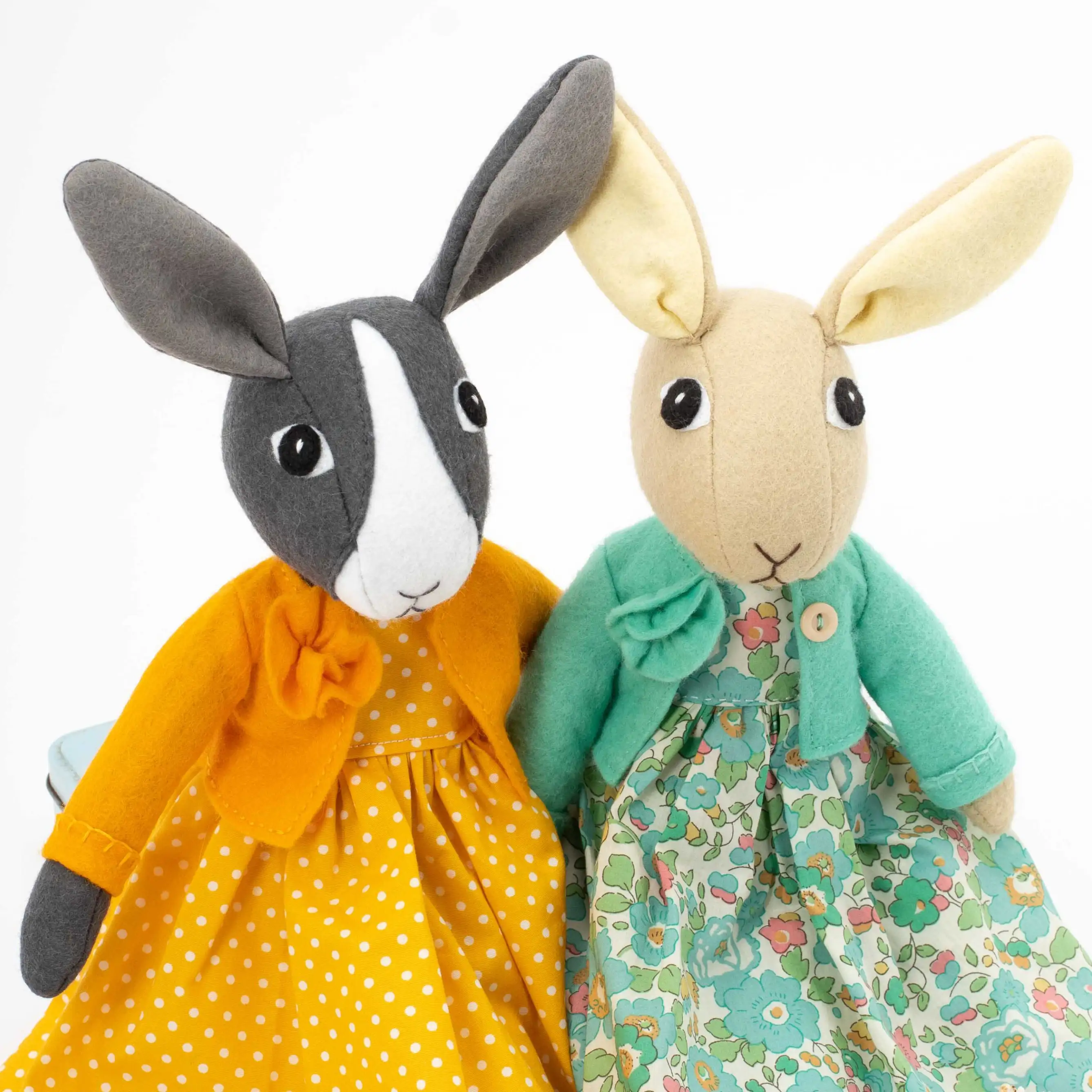 Two felt rabbit toys, one made in light brown felt wearing a green floral Liberty print dress and green felt jacket, and one in grey and white wearing an orange and white polka dot dress with an orange felt jacket.