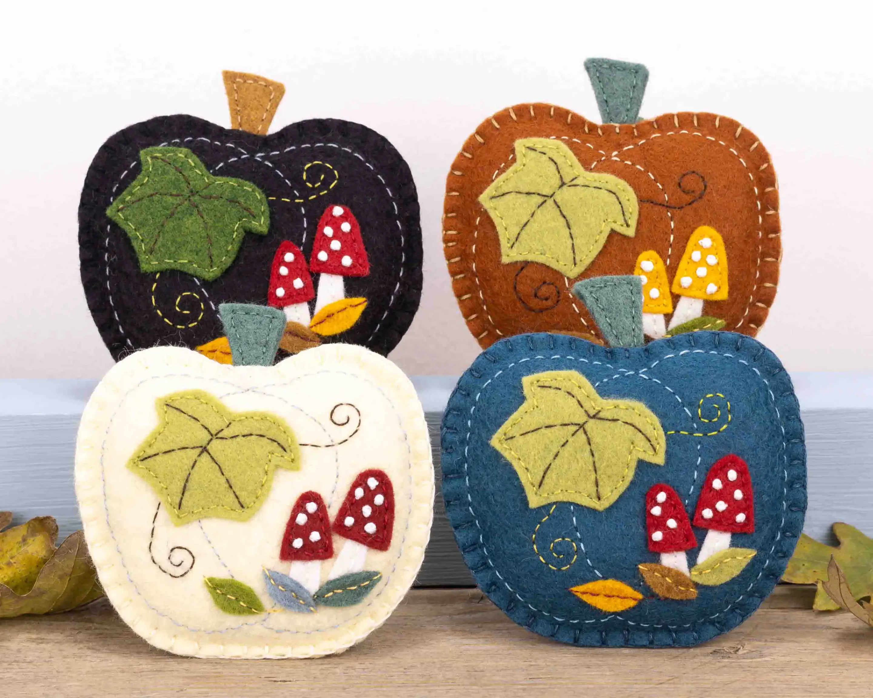 Four pumpkin felt ornaments on a wooden shelf with autumn leaves. Appliqued and embroidered with colourful toadstools and fallen leaves, the pumpkin colours are cream, teal, rust and black.