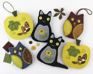 A collection of felt ornaments in warm, bright autumn colours. There are two black cats with yellow button eyes and orange flowers embroidered on them, two yellow-orange pumpkins with leaves and toadstools embroidered on them, and three owls with print fabric wings. Among them are some thread on bobbins, buttons and autumn leaves.