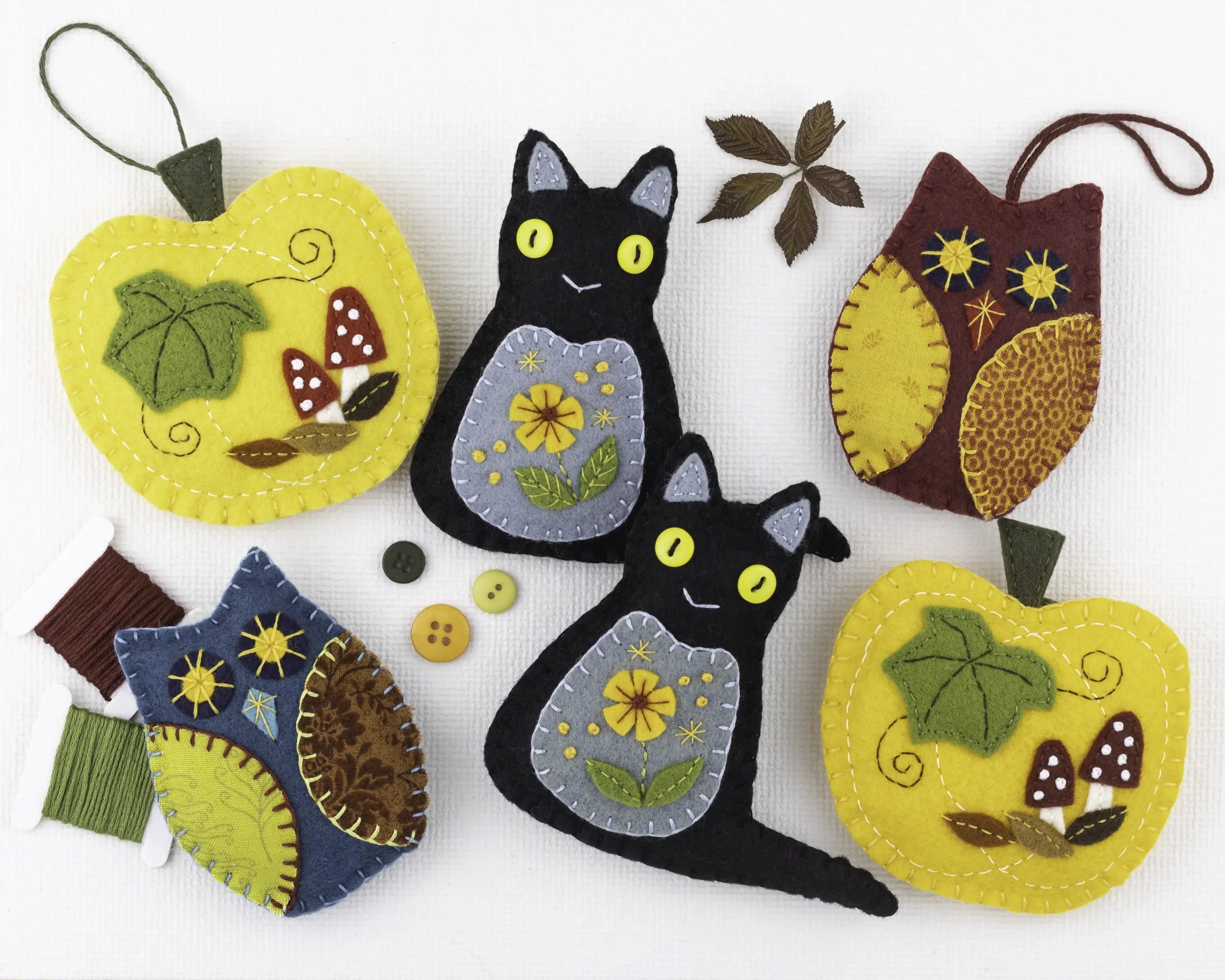 Ornament pattern collection, Autumn A collection of felt ornaments in warm, bright autumn colours. There are two black cats with yellow button eyes and orange flowers embroidered on them, two yellow-orange pumpkins with leaves and toadstools embroidered on them, and three owls with print fabric wings. Among them are some thread on bobbins, buttons and autumn leaves.
