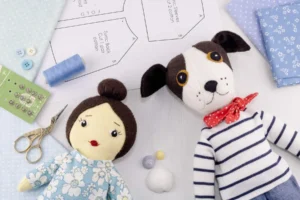 Daisy and Toby dolls lying on a table with blue fabrics, sewing notions and a printed sewing pattern.