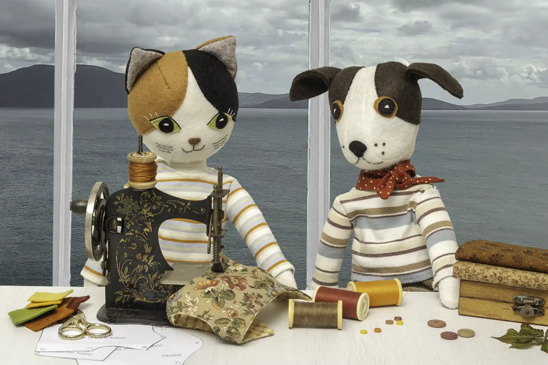 Cat and dog felt dolls sitting at an antique toy sewing machine with autumnal coloured fabrics, threads and buttons. Behind them is a window with a view of the sea and sky.