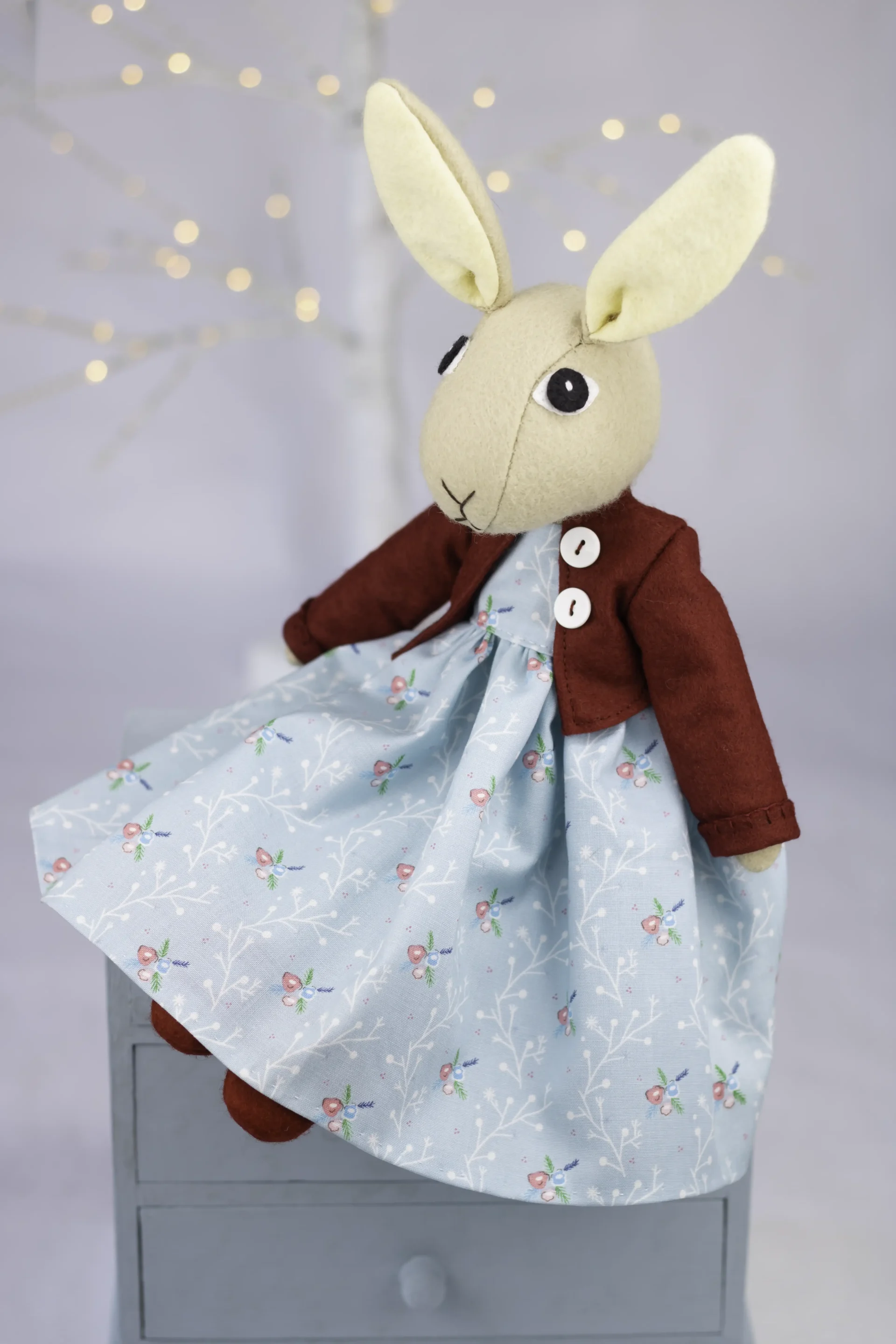 A light brown felt rabbit toy wearing a pale blue floral dress with a long skirt, a burgundy felt jacket with white buttons, and burgundy felt shoes.