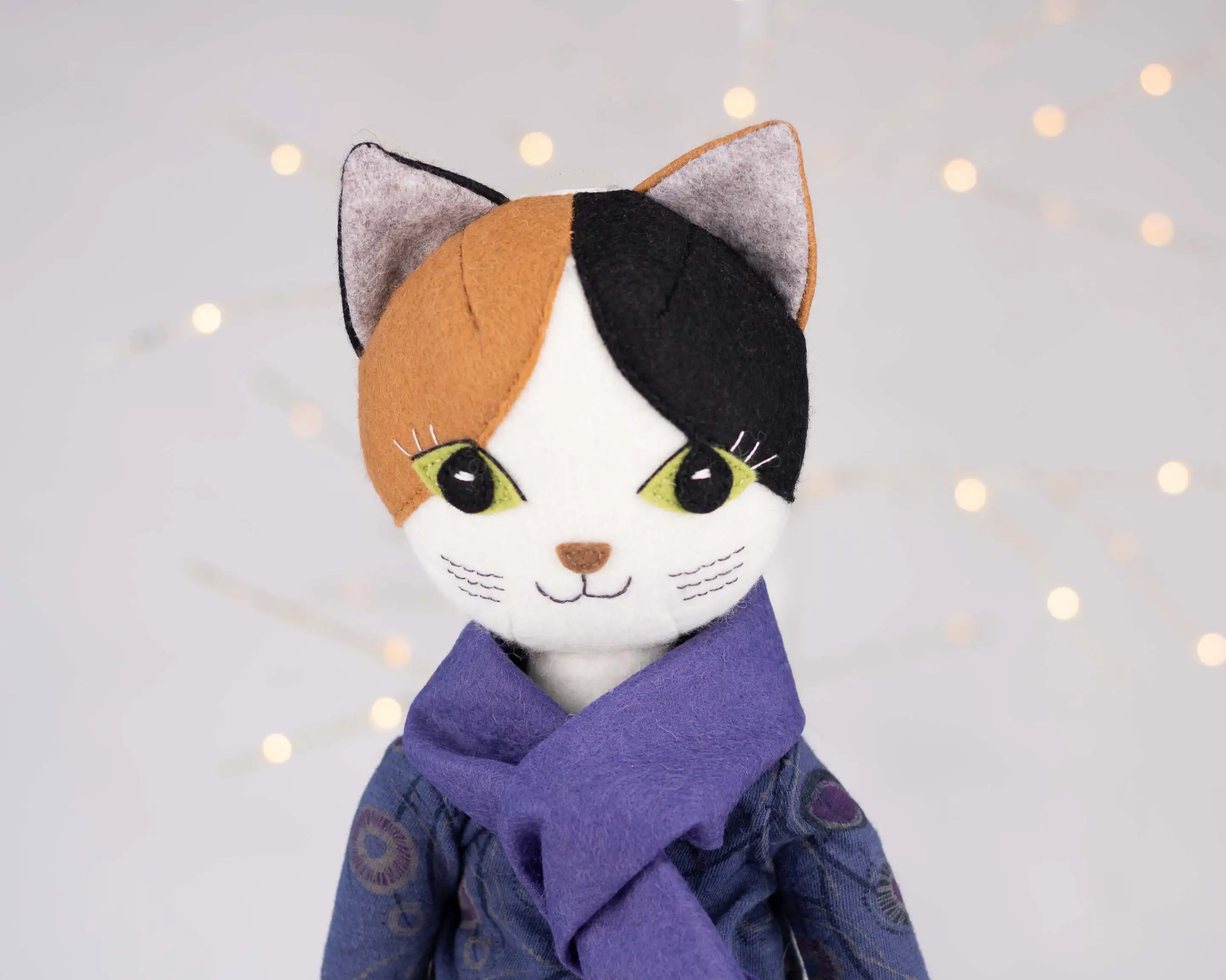 Puffin the cat doll wearing an indigo patterned top and a purple felt scarf, with fairy lights in the background.