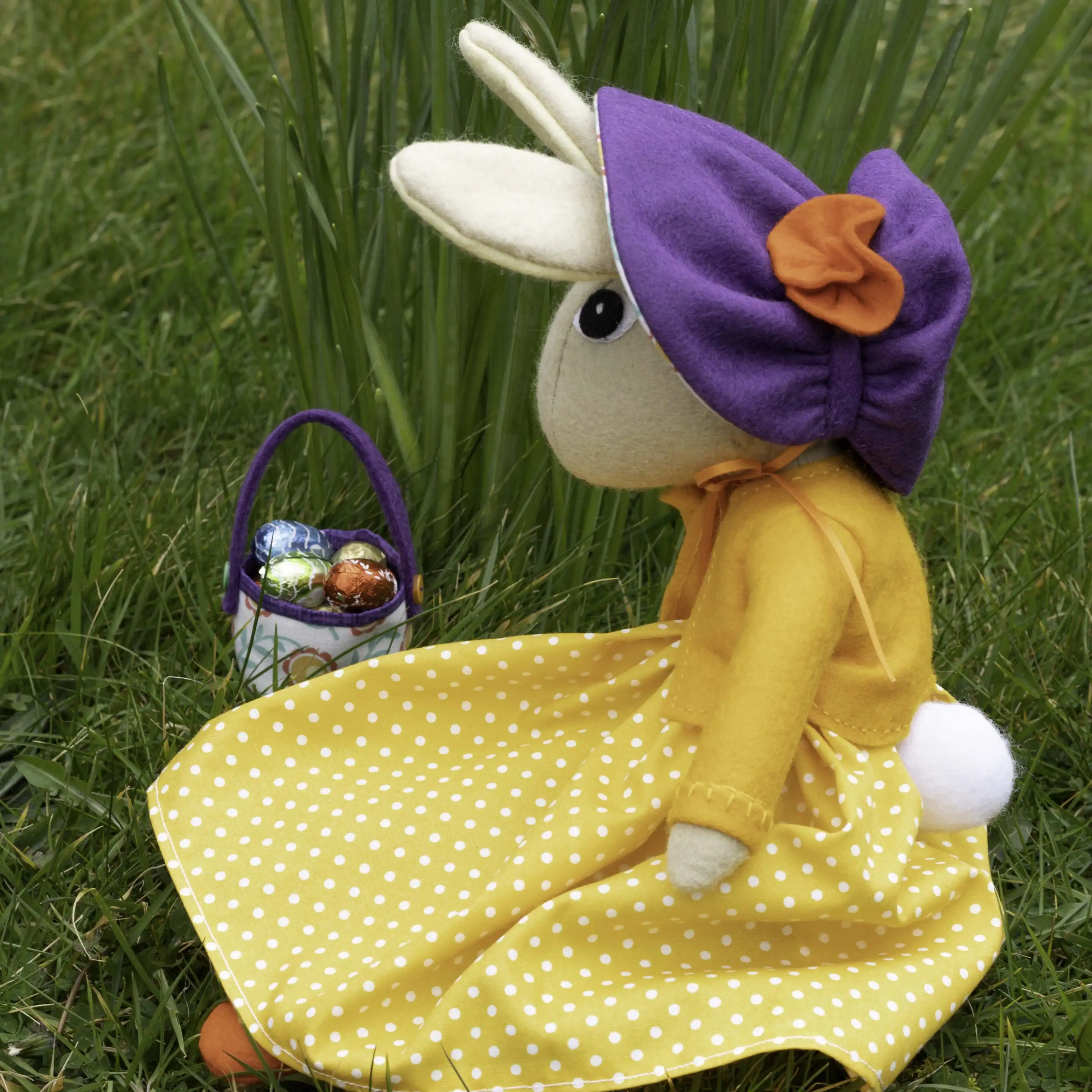A light brown felt rabbit doll wearing an orange and white polka dot dress and orange felt jacket, sitting on grass. She has a purple felt bonnet with an orange flower trim, and a small fabric basket of mini chocolate eggs.