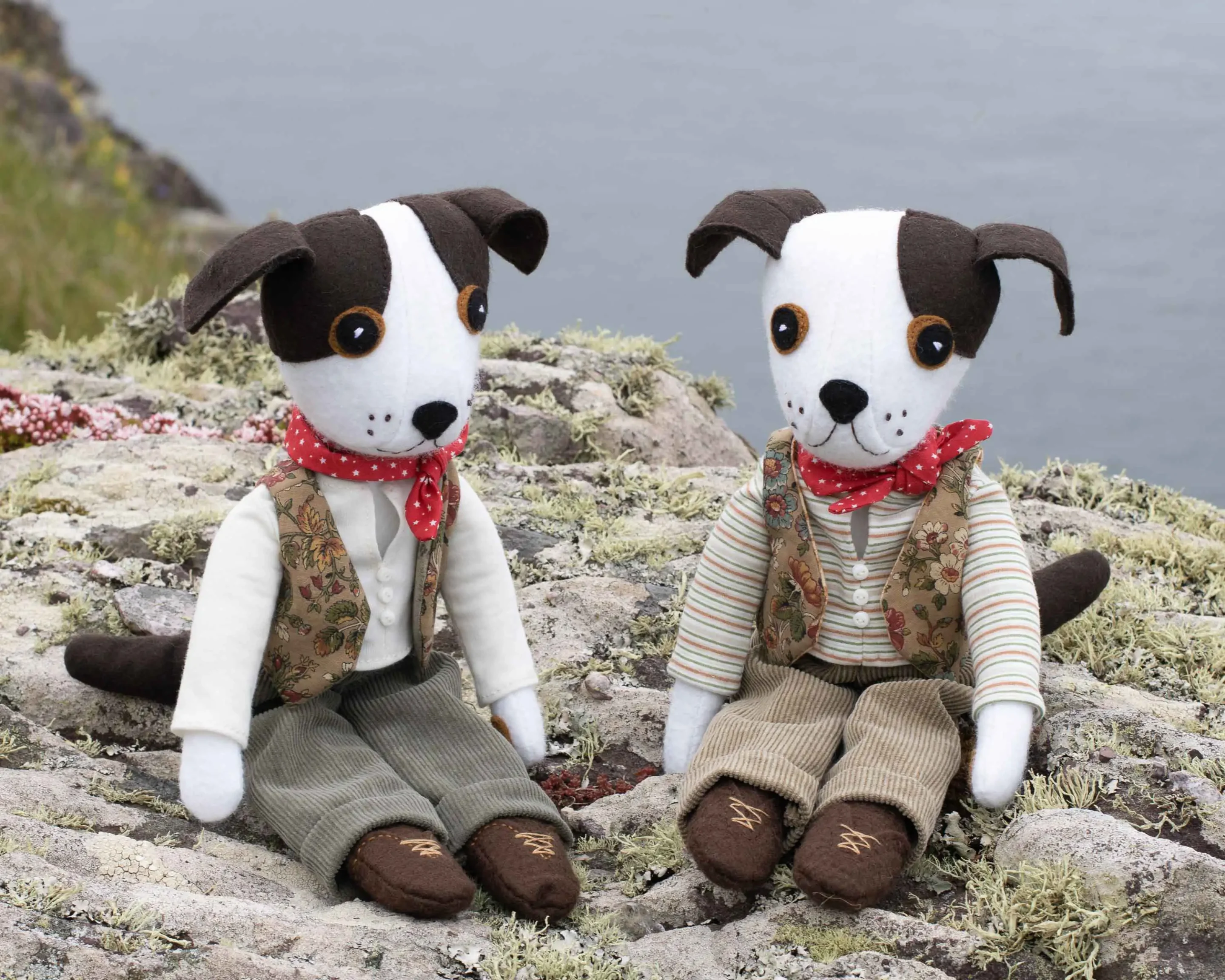 Two Toby dolls sitting on rocks with the sea in the background. One has two brown patches on his face, the other has a single patch.