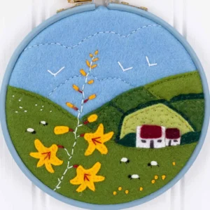 An appliqued and embroidered felt landscape with a farm cottage and sheep, fields in the background and Montbretia flowers in the foreground. The piece is framed in a blue painted wooden embroidery hoop.