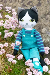 Puffin the black and white cat sits among sea thrift flowers against a lichen covered rock. She is wearing turquoise linen dungarees and a blue and green striped top, with turquoise felt shoes.