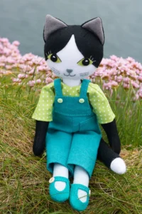 A black and white felt cat doll wearing turquoise linen dungarees and a green and white spot patterned shirt, sitting on a grassy clifftop with sea thrift flowers and the sea in the background.