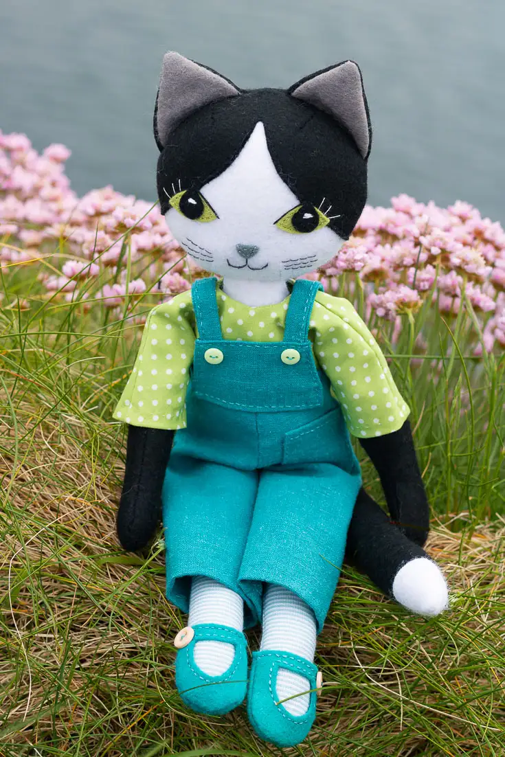 A black and white felt cat doll wearing turquoise linen dungarees and a green and white spot patterned shirt, sitting on a grassy clifftop with sea thrift flowers and the sea in the background.