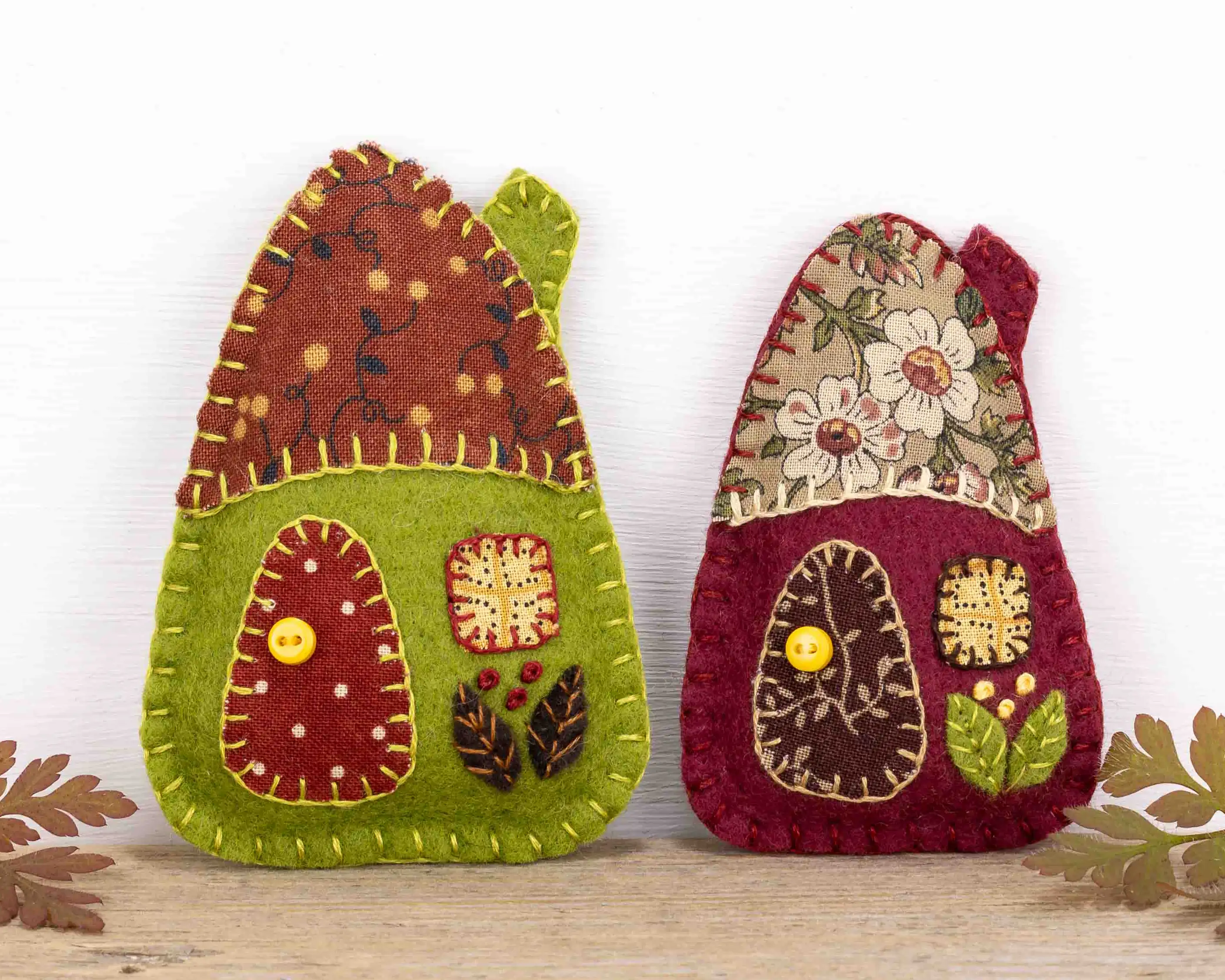 Little felt house ornaments in two sizes, each with a roof, door and window of appliqued patterned fabric. The larger house is olive green and the smaller one burgundy, and they have brown floral roofs. Below the windows are embroidered leaves and berries, and the door knobs are tiny buttons.