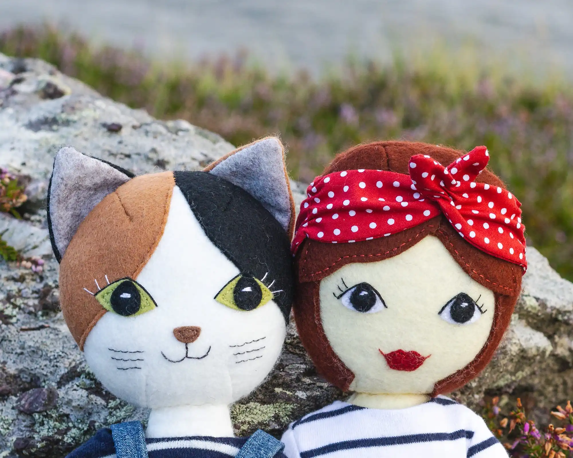 Head and shoulders view of Puffin the Cat and Tilly dolls, sitting against a lichen-covered rock with the sea in the background.