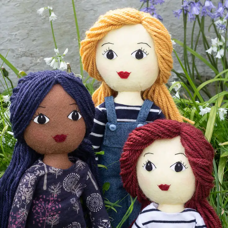 Three felt dolls with long hair made of yarn. One has yellow, one has dark red, and one has dark blue hair. They are outside with bluebells in the background.