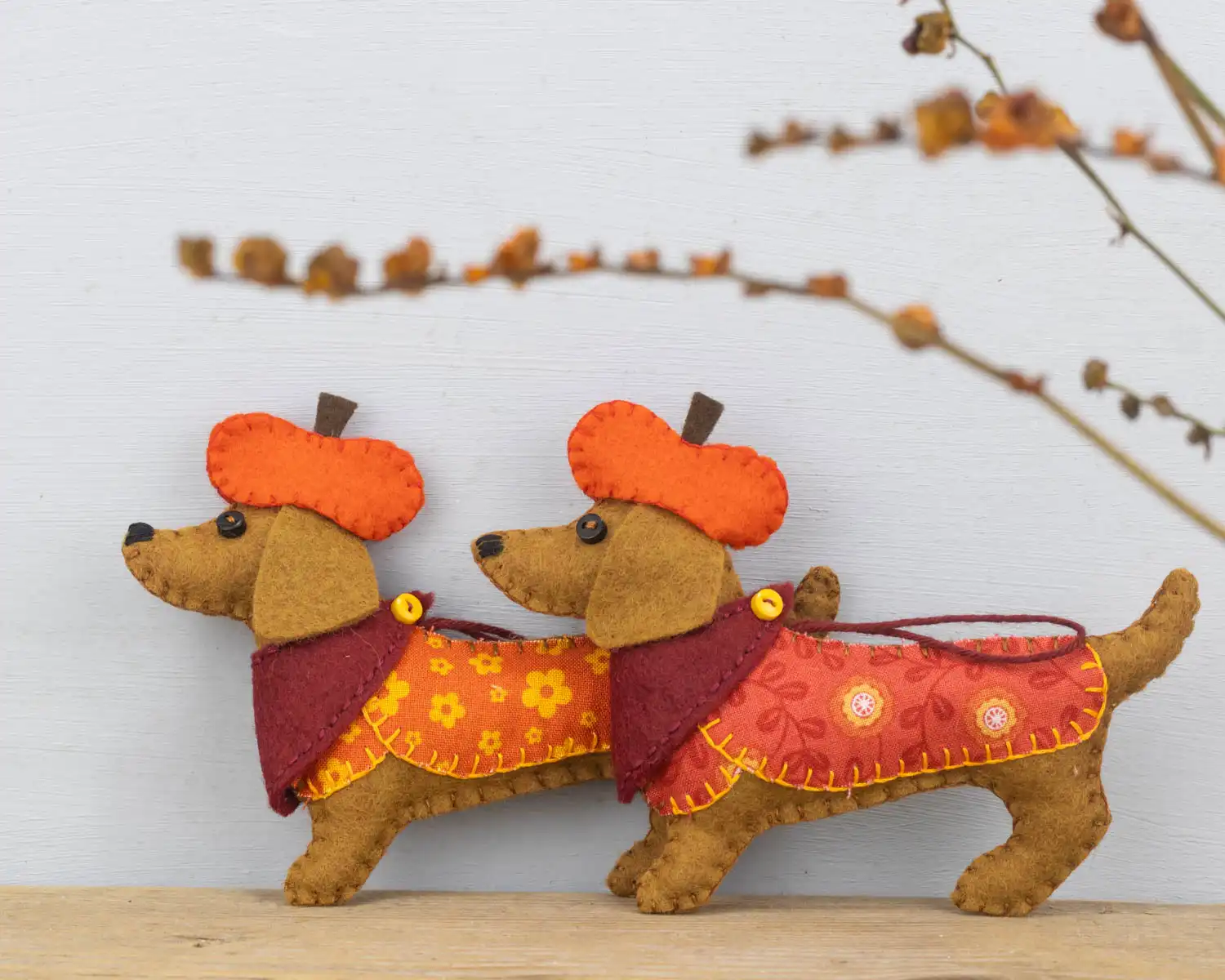 Two Dachshund felt ornaments wearing orange jackets and dark red scarves, with orange pumpkin shaped hats.